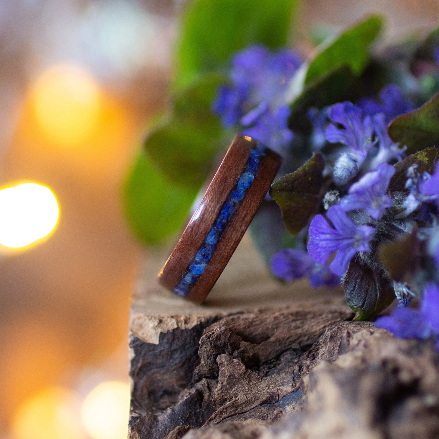 Side view of makore wood ring with striking blue lapis lazuli inlay, propped on bark near flowers.