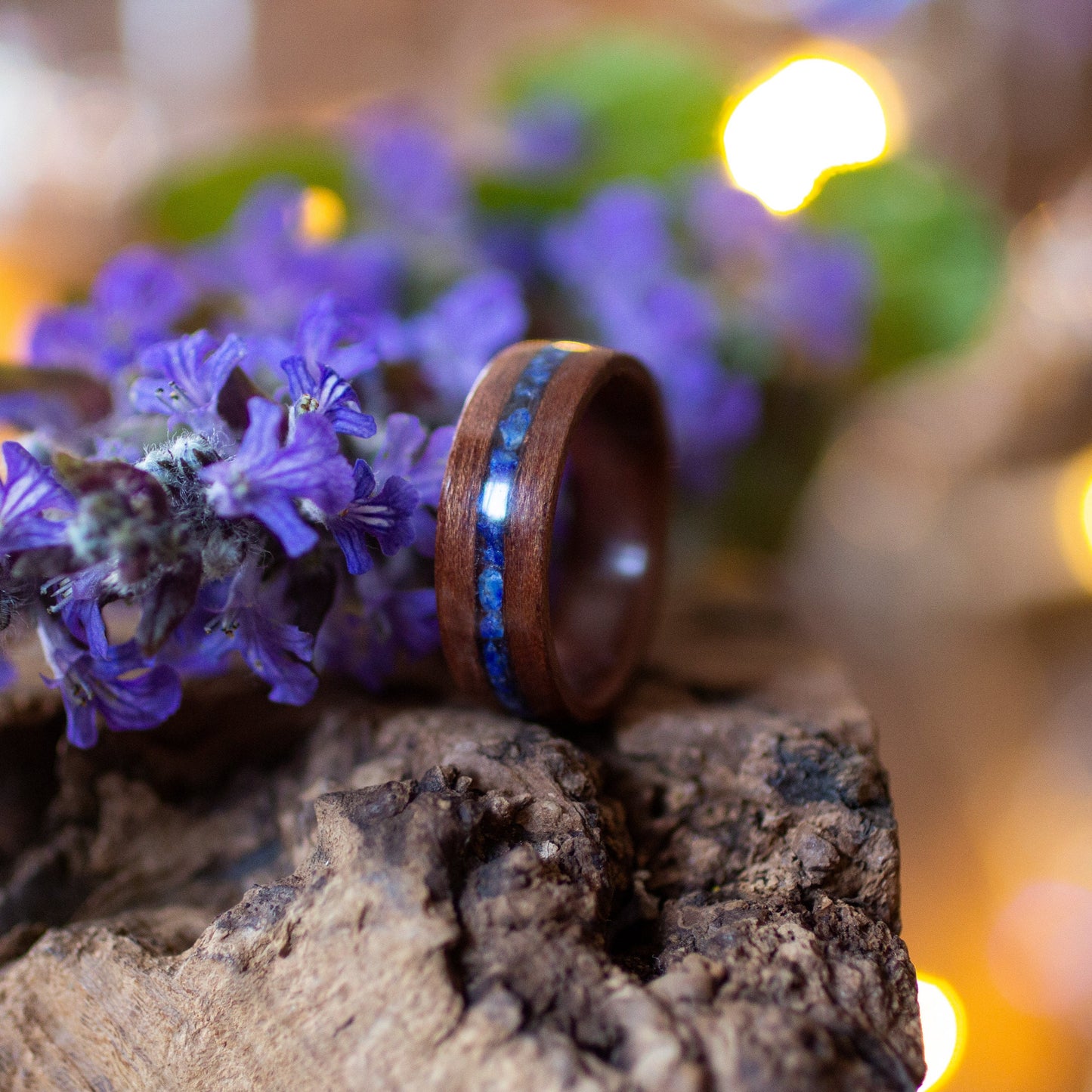 Makore wood ring with lapis inlay beside vivid flowers and greenery, lit by warm ambient light.