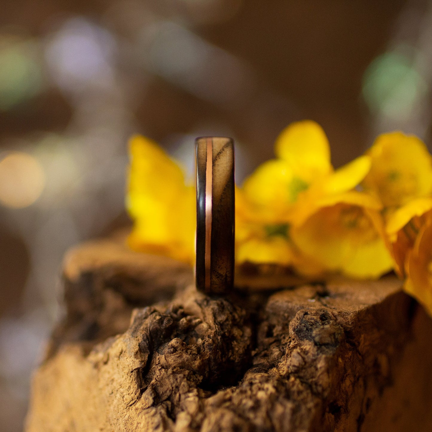 Ash wood ring with copper accents beside yellow flowers on rustic bark, showing grain and polish.