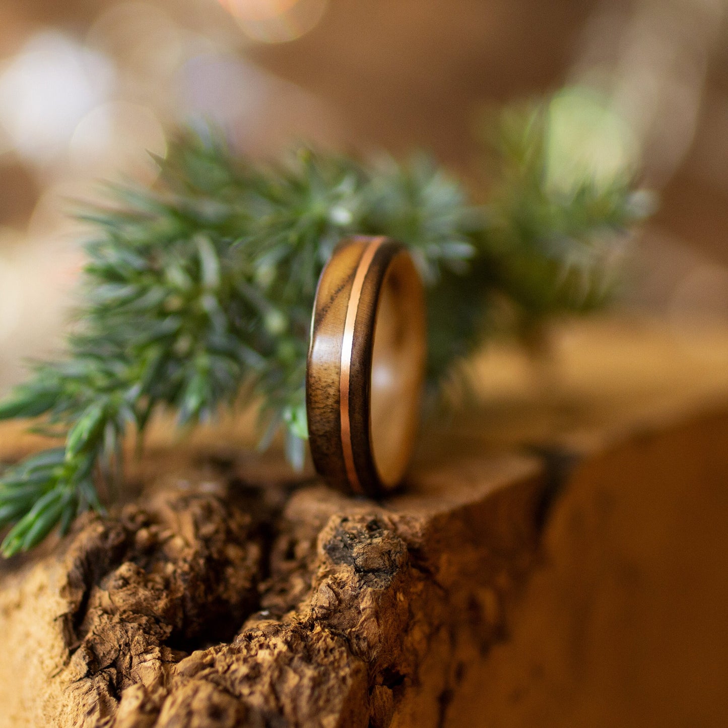 Scorched ash and copper ring styled with pine branch on rugged bark, lit by natural ambient light.
