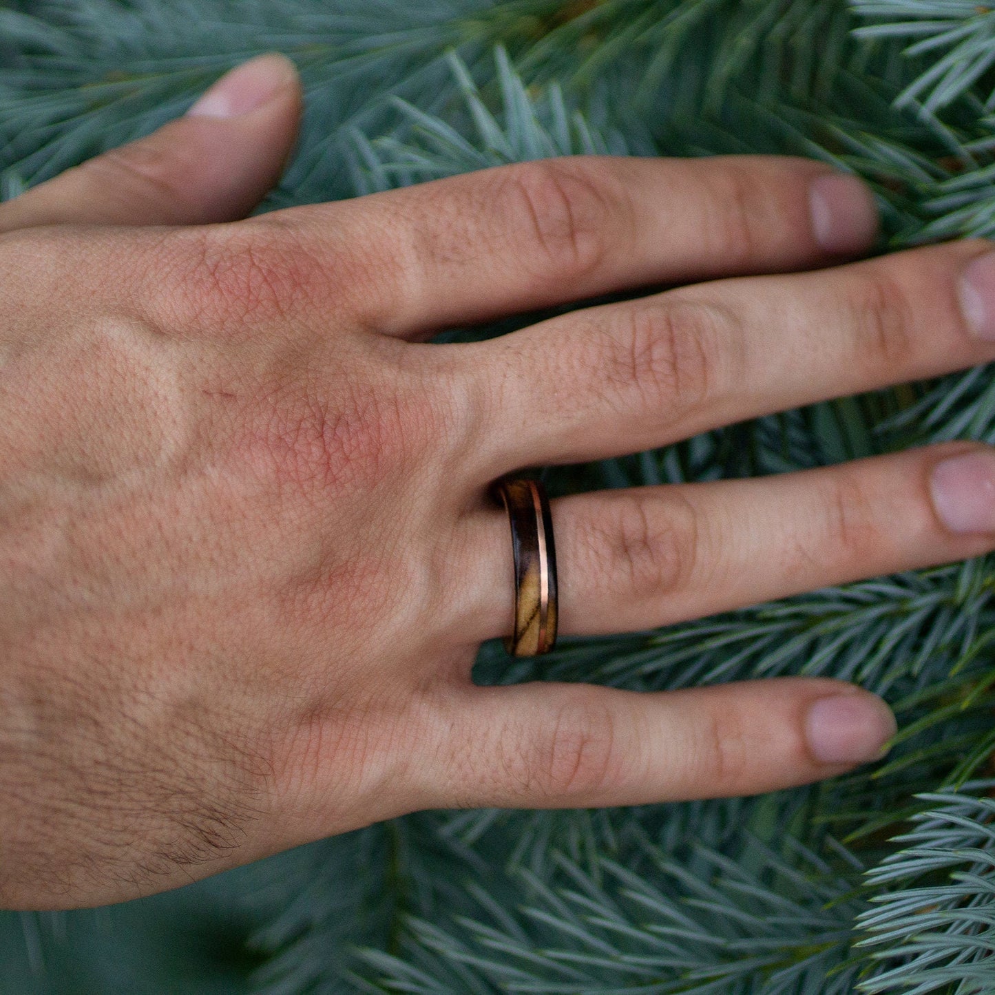 Hand wearing ash wood ring with copper stripe, set against evergreen foliage for natural rustic look.