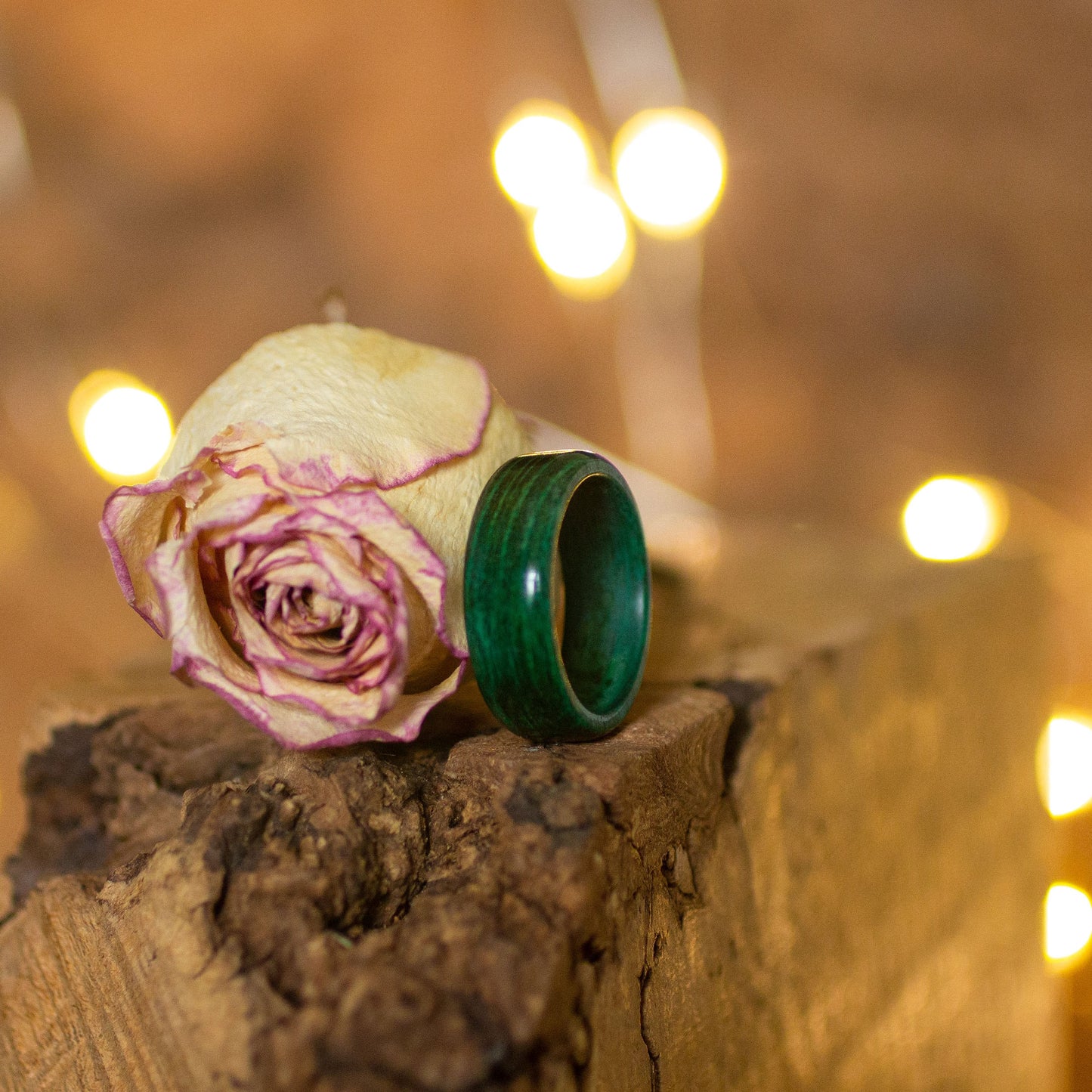 Green-stained wood ring placed near a pale rose, on textured bark with soft bokeh lights.