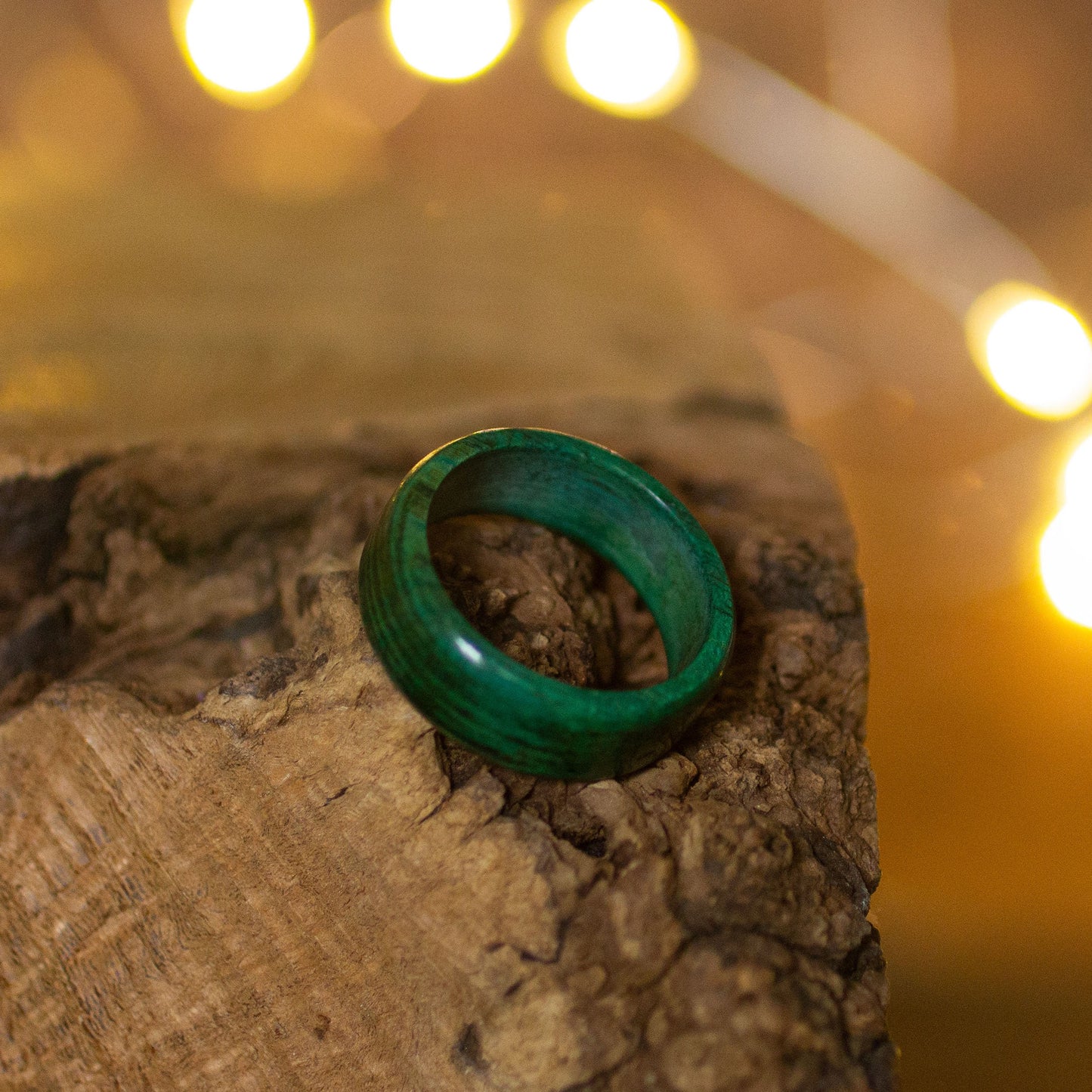 Single green-stained wooden ring displayed on rustic wood grain, catching warm ambient light.