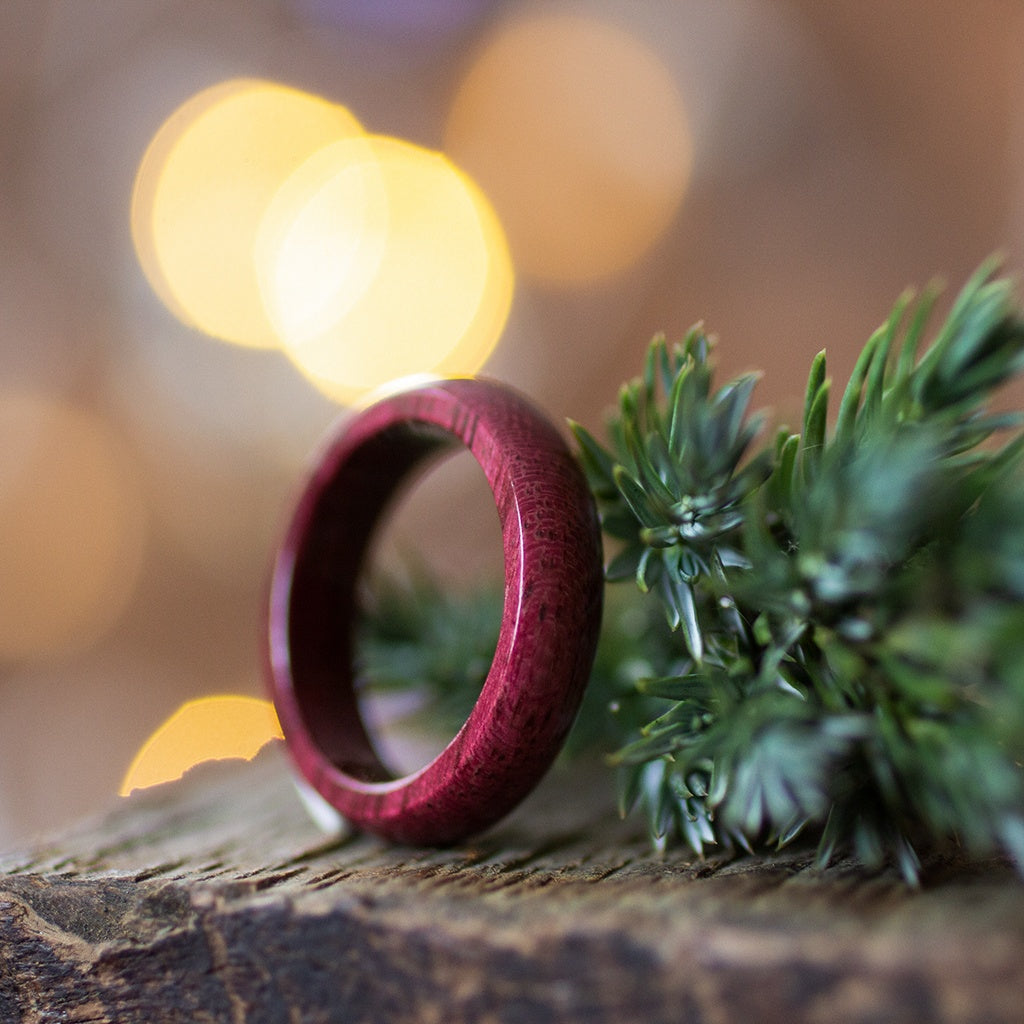 Glossy Purpleheart ring resting on bark with evergreen sprig, warm bokeh lights in the background.