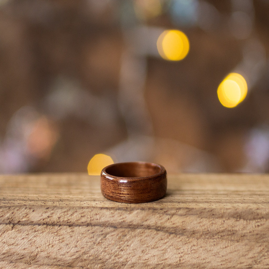 Single makore wood ring with warm reddish tone, lying flat on wood with cozy ambient lights.