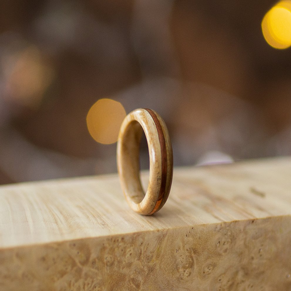 Light ash wood ring with subtle copper detail, standing upright on blonde wood with soft background blur.