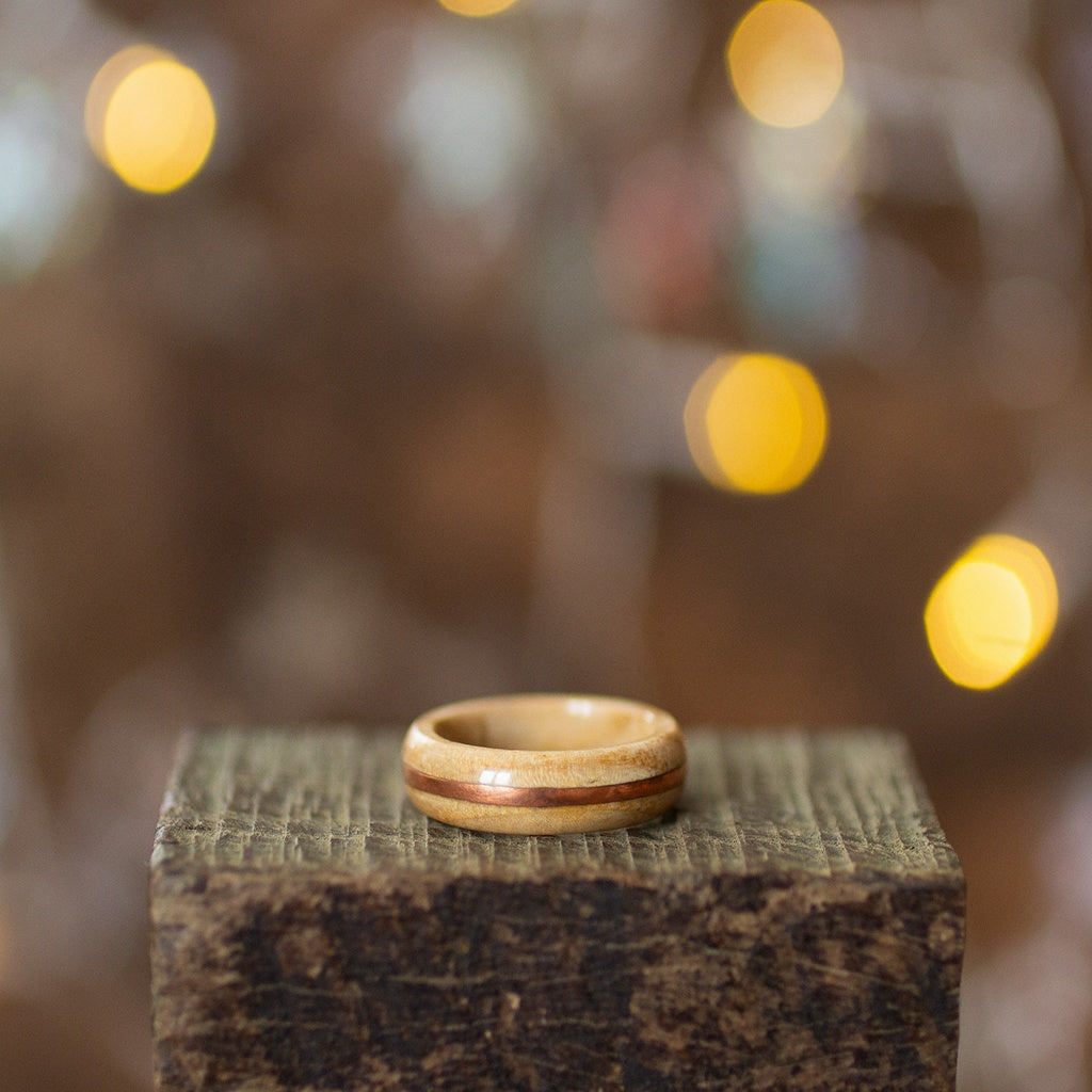 Ash wood ring with warm copper accent, placed on rustic dark wood under warm glowing lights.
