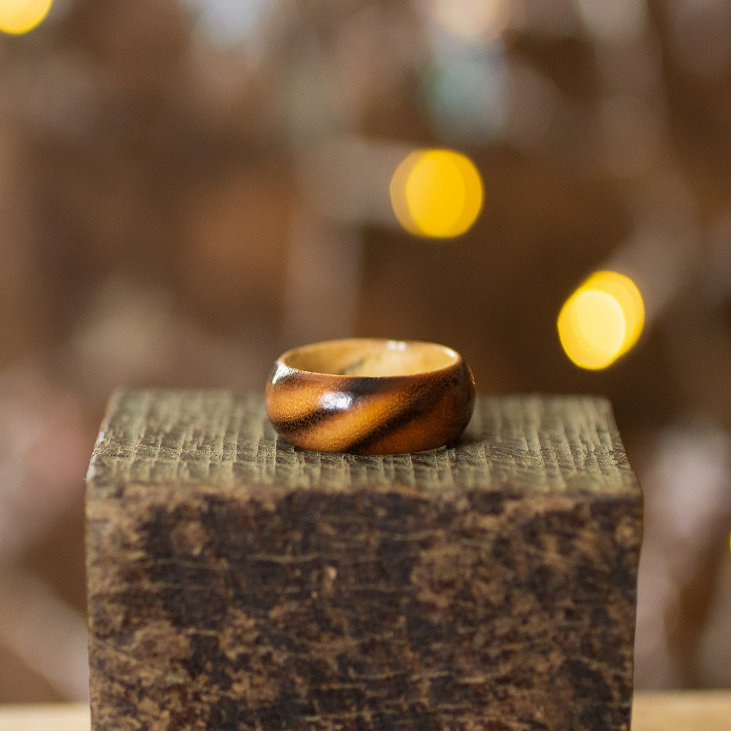 Striped ash ring with two-tone wood grain, displayed on textured wood with golden bokeh background.