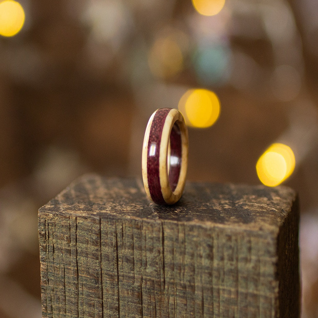 Ash and Purpleheart ring angled to show layered wood grain, with warm ambient lights in background.