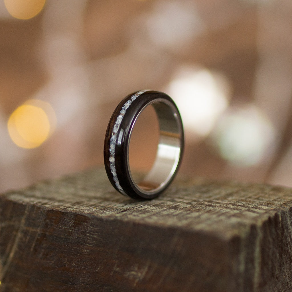 Ebony wood ring with crushed pearl inlay, standing upright on rustic wood with soft bokeh lights.