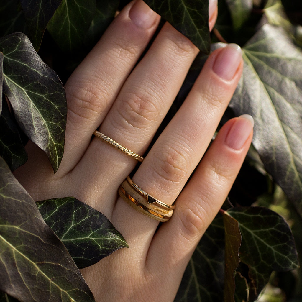 Close-up of hand wearing light ash ring with copper inlay and stacking gold ring, framed by greenery.