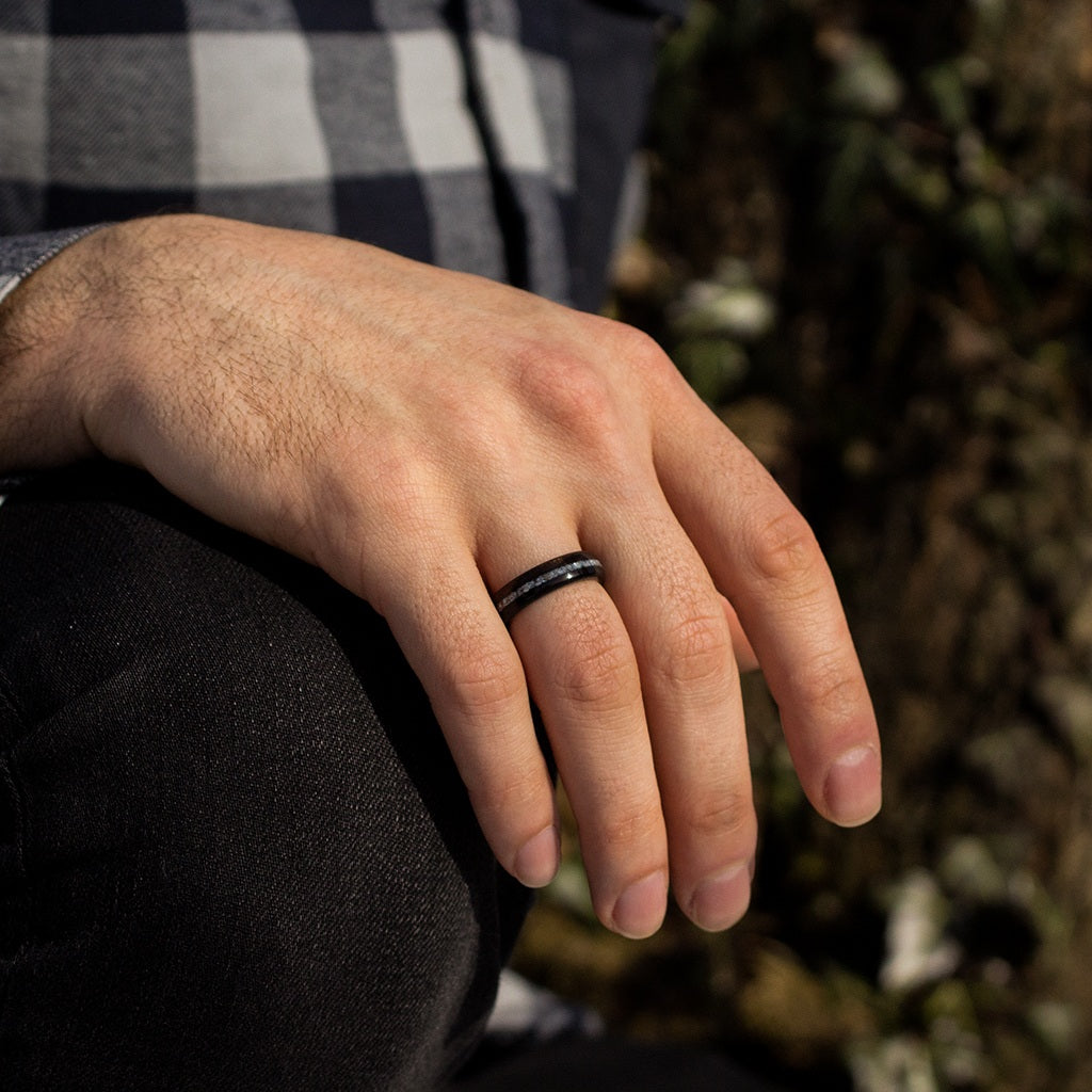 Man’s hand wearing ebony wood ring with pearl inlay, resting on knee with outdoor background.