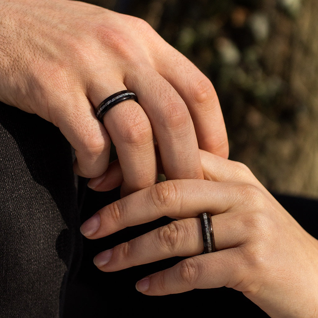 Close-up of couple holding hands, both wearing ebony rings with pearl inlay, soft light and earthy tone.
