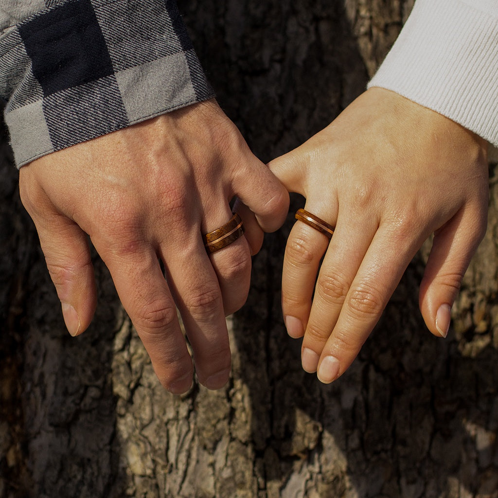 Two hands with mahogany and copper rings placed on tree bark, natural outdoor setting.