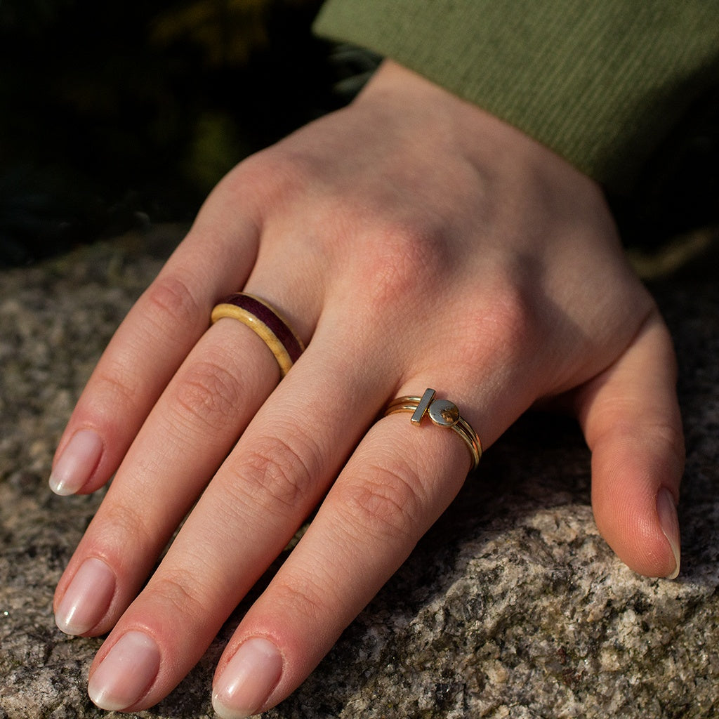 Close-up of hand wearing ash and Purpleheart ring, resting on stone with natural textures.