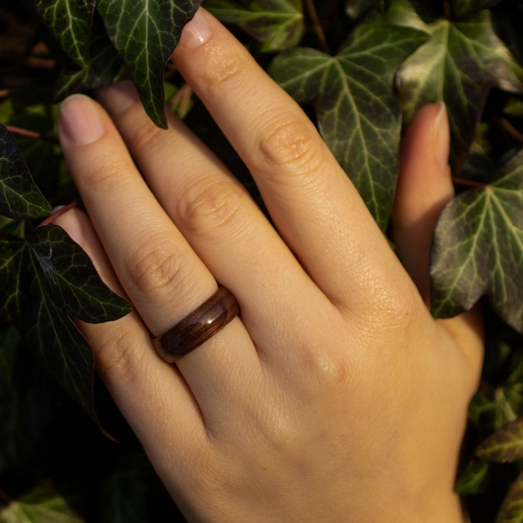 Makore wood ring worn on middle finger, hand resting on tree trunk with foliage behind.