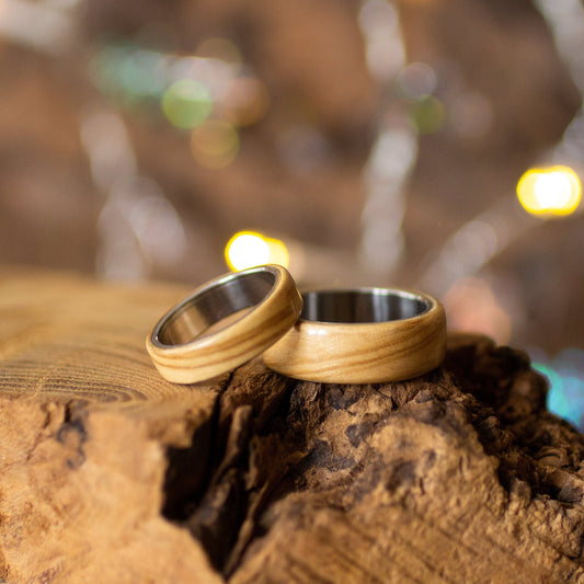 Pair of olive wood rings with stainless steel core resting on textured bark, softly lit with warm bokeh.