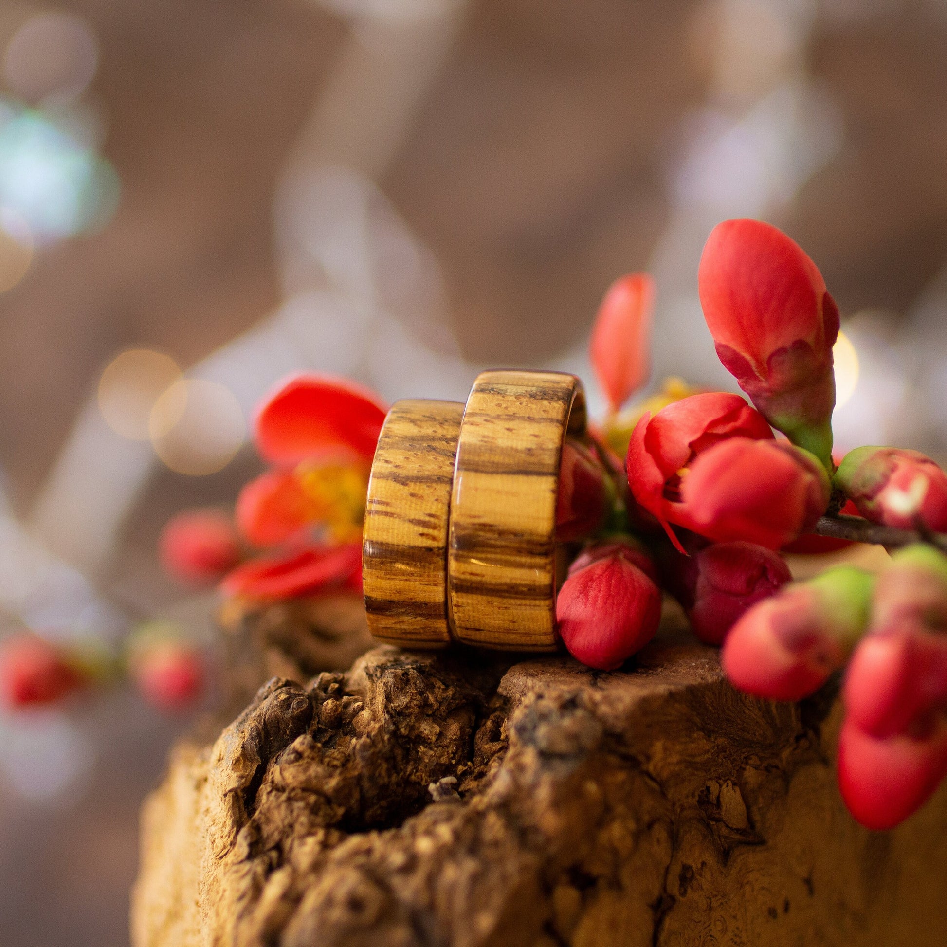 Side view of Zebrano rings with bold striped grain, resting on rustic bark with floral accents.