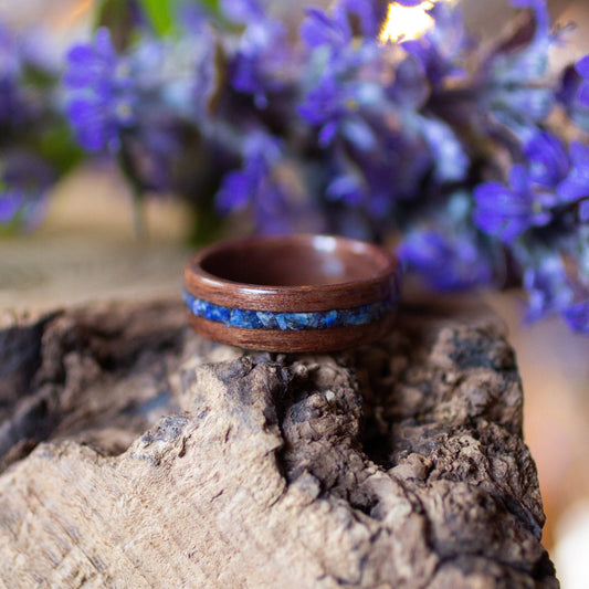 Makore wood ring with deep blue lapis lazuli inlay, displayed on bark with soft-focus purple flowers.