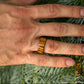 Hand wearing Zebrano bentwood ring, showing striped wood grain against evergreen branches.