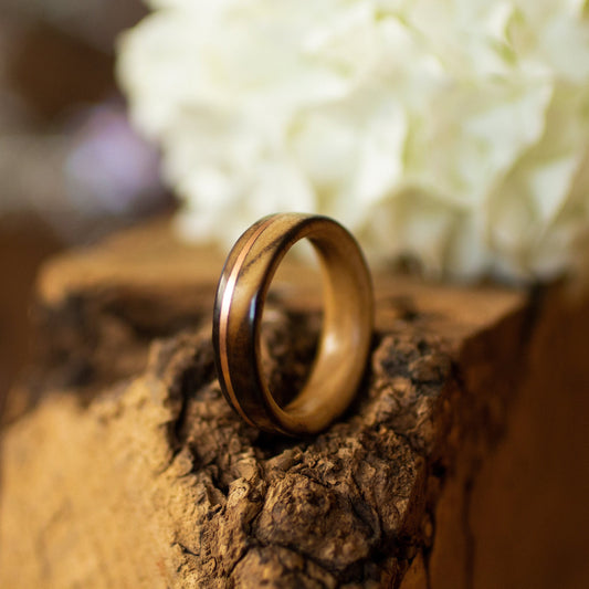 Scorched ash wood ring with copper inlay, standing on textured bark with white hydrangea in background.