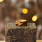 Striped ash ring with two-tone wood grain, displayed on textured wood with golden bokeh background.