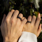 Couple’s hands wearing matching ebony and pearl rings, surrounded by ivy leaves and natural light.