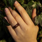 Makore wood ring worn on middle finger, hand resting on tree trunk with foliage behind.