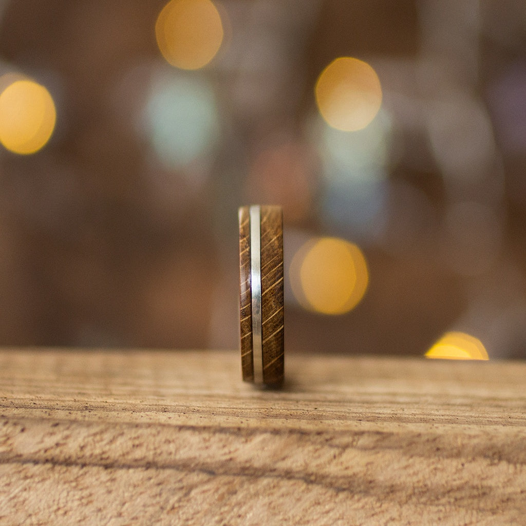 Vertical view of recycled oak wood ring with thin silver inlay, standing on light wooden surface with blurred warm lights.