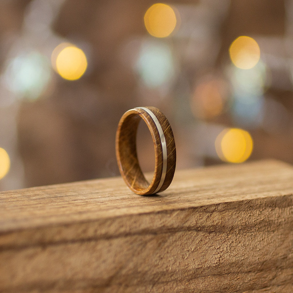Handmade oak ring with silver inlay, captured from rear angle on rustic wood, warm golden bokeh behind.
