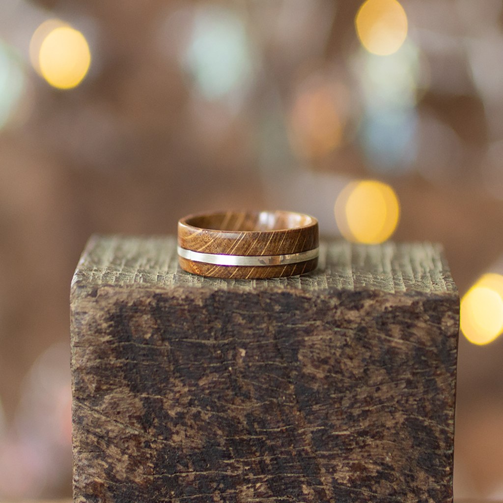 Recycled oak wood ring with silver inlay by Andrwood, placed flat on rustic block, softly lit with bokeh in background.